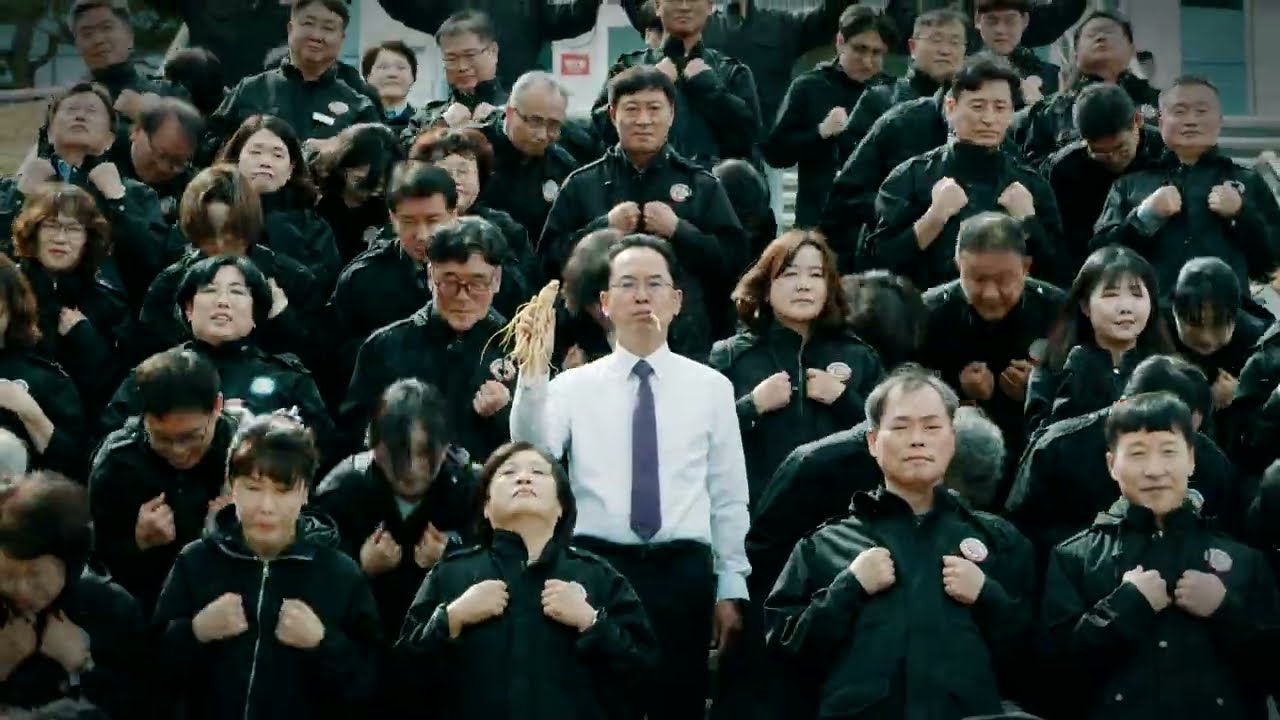 高麗人参を手に中央に立つ韓国・栄州市の副市長と、その背後の階段で一斉に踊る黒い制服姿の幹部職員数十人